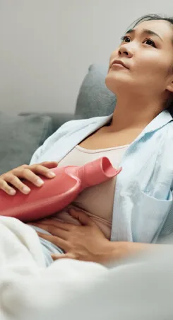 A woman lying on a couch holding a hot water bottle against her abdomen.