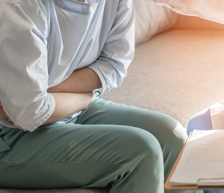 A woman sitting on a bed holding her abdomen in discomfort.