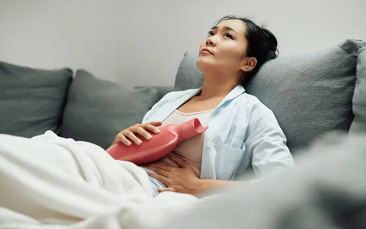 A woman lying on a couch holding a hot water bottle against her abdomen.